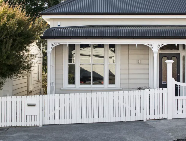Street-facing view of a traditional single-storey residential facade with pale horizontal weatherboard cladding, ornate white veranda fretwork and posts, a corrugated dark metal roof and matching veranda awning, white picket fencing, and a dark timber front door. The image focuses on the exterior architecture and frontage.