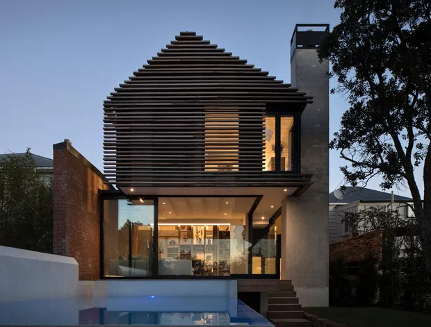 A contemporary multi-level urban residence photographed at dusk, with a strongly sculptural gabled roof and upper facade screened by closely spaced horizontal timber slats. The lower level has extensive floor-to-ceiling glazing opening toward a reflecting pool, while exposed brick, concrete-rendered walls, dark window frames, and warm interior lighting create a refined, dramatic composition. Mature trees and neighbouring homes frame the site.