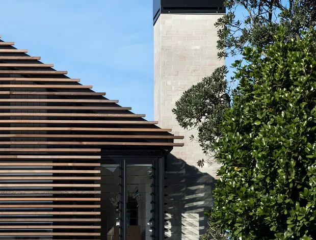 Exterior view of a contemporary multi-level residential building. The facade combines extensive horizontal timber slat screening with dark-framed glazing and pale concrete masonry walls. A tall rectangular tower with a dark parapet and rooftop cylindrical vent rises behind the slatted facade, while mature landscaping partially obscures the building. Bright clear daylight emphasizes the strong geometric lines and layered facade.