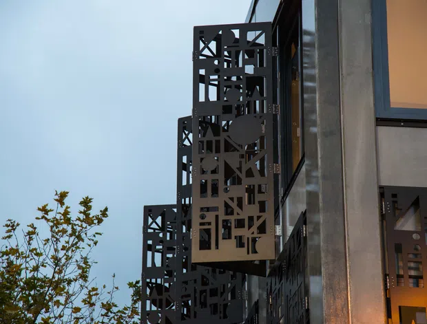 Exterior architectural detail viewed upward along a contemporary multi-storey facade. Dark perforated metal screens with abstract geometric cut-outs project from the building, alongside smooth grey wall panels, dark-framed glazing, and warm interior light visible through openings. A small tree and pale sky occupy the background.