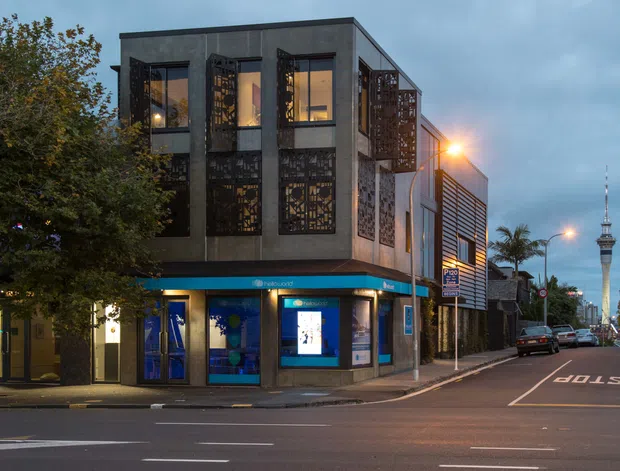Street-level dusk view of a contemporary multi-storey commercial building on an urban corner. The dark rendered facade has large windows and extensive perforated decorative metal screens, with illuminated retail frontage at ground level. A marked city street, neighbouring buildings and the Sky Tower are visible in the background.