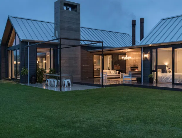A low, modern residential house photographed from the lawn at dusk. The building has multiple pitched metal-roofed wings, dark vertical cladding, extensive floor-to-ceiling glazing, a prominent tall masonry chimney, and a covered outdoor dining terrace with a slim dark frame. Warm interior lighting reveals an open living/dining area and a bedroom through the glass. The house sits in a spacious grassy setting with distant wooded hills.