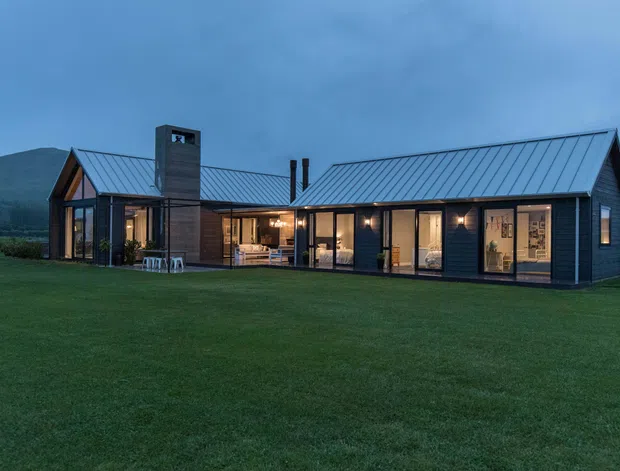 A low, elongated contemporary house photographed at dusk, set in an expansive grassy rural or alpine landscape. The dark horizontal-clad facade combines extensive floor-to-ceiling glazing with a steep gabled roof and a projecting glazed living space at the left. A prominent masonry chimney rises through the roof, while warm interior lighting creates a welcoming glow against the cool blue exterior light.
