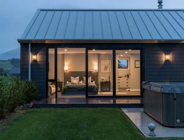 Exterior view of a compact dark-clad dwelling at dusk, featuring a standing-seam metal roof, full-height glazed doors, and warm interior lighting. A bedroom and bathroom/vanity area are visible through the glazing, with a spa or hot tub on the paved terrace in the foreground and a landscaped lawn surrounding the building. Mountainous rural scenery is visible in the distance.