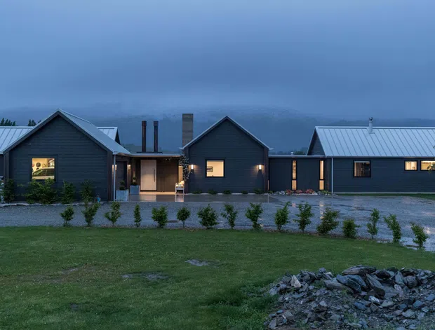 Exterior view of a contemporary single-storey residence composed of several connected gabled volumes. The building has dark horizontal cladding, bright standing-seam metal roofs, warm illuminated windows, and a recessed central entry. It is set in a landscaped rural-looking site with gravel paths and low mountains or hills visible through the dusk haze.