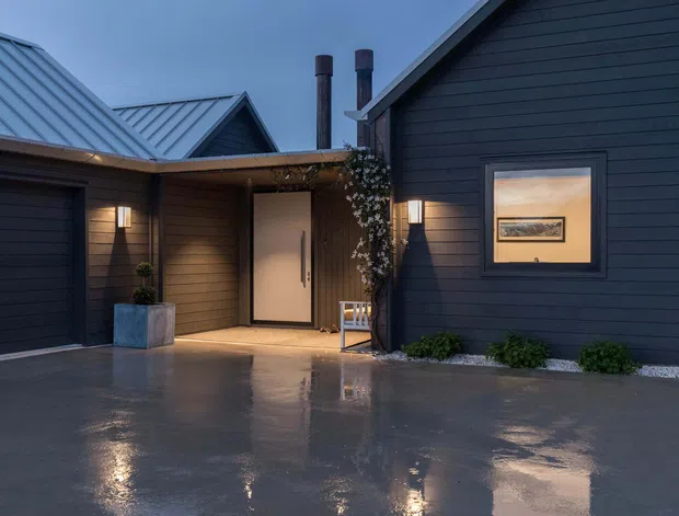 A dark, contemporary residential entrance courtyard photographed in wet evening conditions. The composition shows charcoal horizontal cladding, a covered entry with a pale pivot door, a window with warm interior light, metal pitched roof sections, exterior wall lights, restrained planting, and a reflective paved driveway or forecourt.