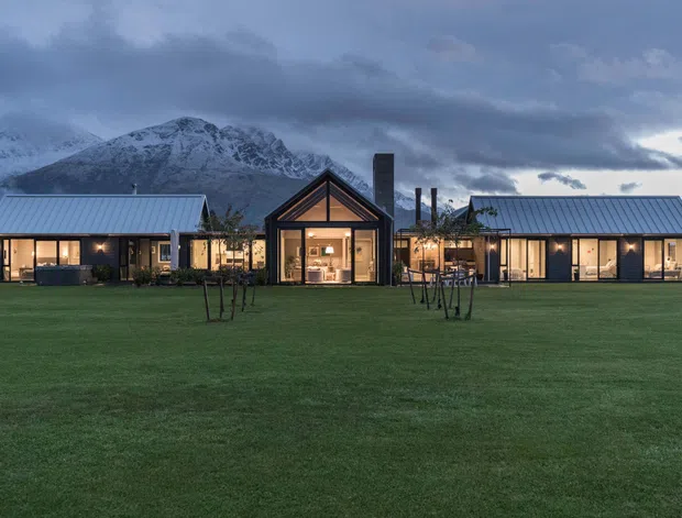Wide exterior view of a low-rise residential property arranged as three connected or closely grouped pavilion-like forms across a manicured lawn. The buildings feature dark vertical cladding, large glazed openings, simple pitched metal roofs, and a prominent central gabled volume with warm interior lighting. Snow-covered mountains and overcast dusk skies form the background, while young trees and landscaped grass occupy the foreground.