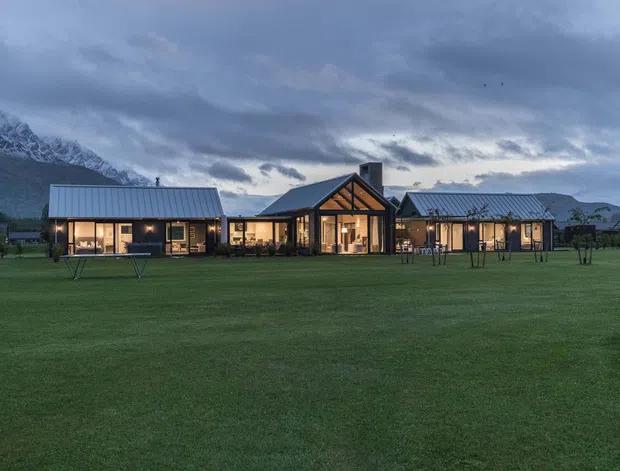 A wide exterior view of a low-slung, single-storey building set within a large manicured lawn, with dramatic snow-capped mountains and overcast skies behind it. The building has multiple connected wings with dark pitched metal roofs, extensive warm-lit glazing, timber-framed gable ends, and a contemporary lodge-like form.