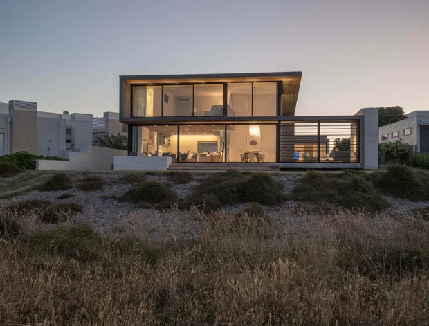 Exterior view of a contemporary two-storey residence at dusk, featuring a strong rectangular composition, extensive floor-to-ceiling glazing, dark horizontal framing, a projecting upper volume, and a covered open-sided structure to the right. The house sits behind low coastal-style vegetation and landscaped ground, with warmly lit interiors visible through the windows.