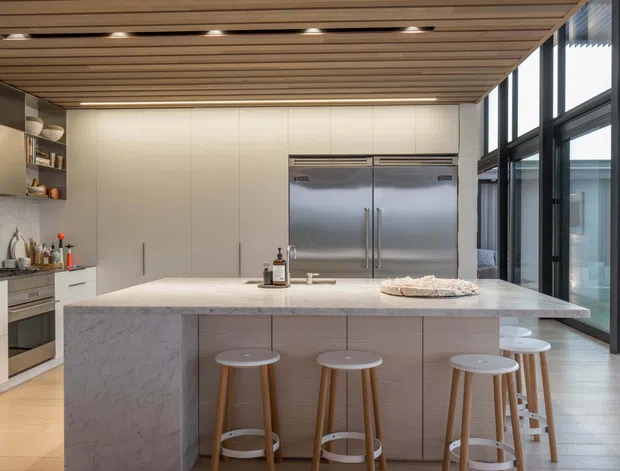 A bright, spacious contemporary kitchen centered on a large marble-look island with seating for five or more. The kitchen features pale fixed cabinetry, integrated appliances, a stainless-steel double-door refrigerator, expansive black-framed glazing, and a timber slatted ceiling with recessed lighting. The palette is light and neutral with warm timber accents.