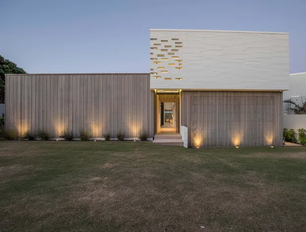 Front exterior view of a contemporary single-storey residence at dusk. The facade combines vertically ribbed timber-clad volumes with a clean white rendered upper box featuring a patterned perforated detail, warm recessed entry lighting, and simple landscaped lawn frontage.