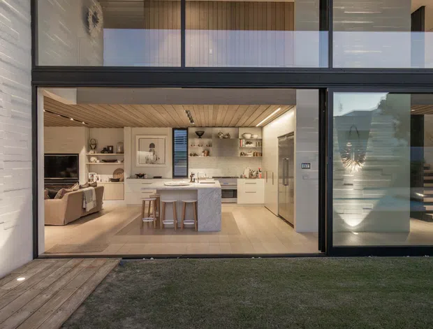 View from a small landscaped courtyard into a contemporary open-plan kitchen and living area through expansive black-framed glazing. The interior features a central stone island with stools, pale built-in cabinetry, integrated shelving, timber flooring, and a warm timber-lined ceiling. A glazed upper level and a staircase are visible, emphasizing the home's modern, open composition.