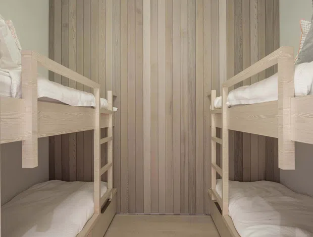 Compact bunk-bed room with two sets of built-in timber bunk beds facing each other. The walls are clad in vertical natural timber boards, with pale timber bed frames, integrated ladders, white bedding, and a simple timber floor. The symmetrical layout and enclosed scale suggest a practical children’s or shared sleeping space.