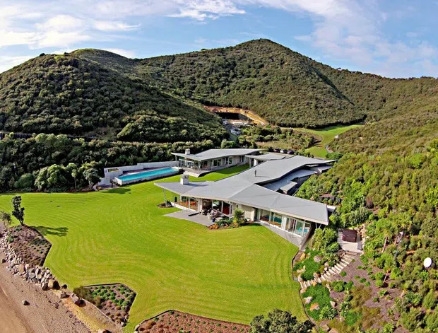 Aerial view of a large contemporary residence set within a dramatic green coastal or bush-covered hillside landscape. The sprawling low-pitched roof forms surround landscaped courtyards and terraces, with expansive glazing, a long turquoise swimming pool, manicured lawns, planted garden beds, and a rocky shoreline visible at the edge of the property.