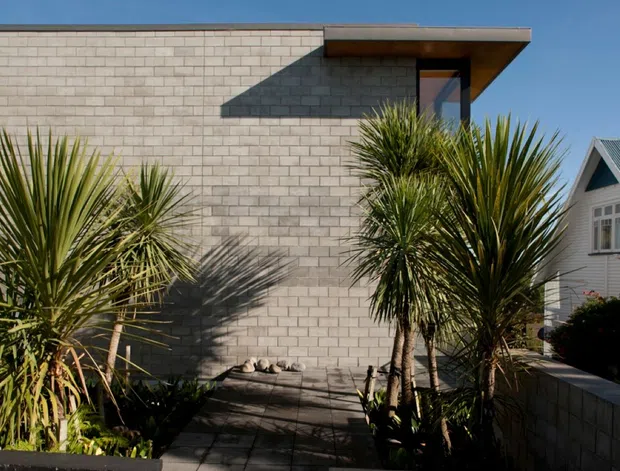 Exterior architectural photograph of a contemporary single-storey building with a flat roof, deep projecting eave, narrow vertical window, and pale grey concrete block walls. A paved walkway and mature spiky planting frame the facade, with a traditional gabled house visible in the background.