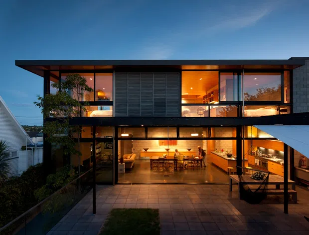 A contemporary multi-level residential house photographed at dusk, with extensive floor-to-ceiling glazing revealing warmly lit living and dining areas. The upper level features dark horizontal screening and timber-toned interiors, while the lower level opens onto a paved courtyard or terrace. Black structural framing, a flat roofline, and a projecting white roof or canopy create a strong modern composition. Neighbouring homes and limited landscaping are visible around the property.