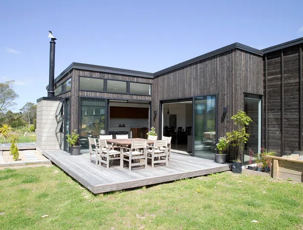 Exterior view of a contemporary single-storey home with dark vertical timber cladding, extensive glazing, a broad low-pitched roofline, and a timber deck furnished with an outdoor dining table and chairs. A chimney and light masonry feature wall are visible, with landscaped lawn surrounding the building.