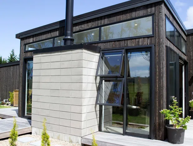 Exterior view of a contemporary single-storey building with dark vertical timber cladding, extensive black-framed glazing, and a projecting light masonry-clad chimney volume with a tall black flue. A raised timber deck, gravel garden beds, stepping stones, and small planted shrubs form the foreground.