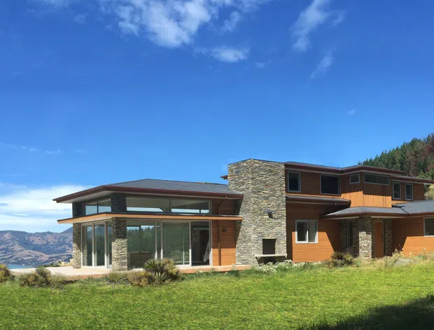 A contemporary multi-level hillside residence with extensive floor-to-ceiling glazing, warm timber-clad walls, a prominent natural stone chimney or feature wall, and broad low-pitched roof planes. The house sits in a grassy elevated landscape with rugged hills and a body of water visible in the distance.