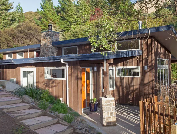 Exterior view of a compact, low-rise home set into a wooded, sloping site. The building features dark vertical timber cladding, broad low-pitched roof planes, extensive horizontal glazing, and a prominent rough-stone chimney. Stepping-stone paving leads to the entrance, with timber screening and planting around the front.
