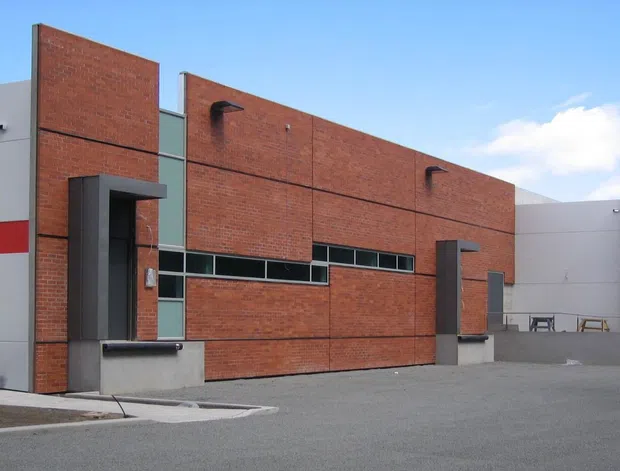 Exterior view of a contemporary institutional or commercial building composed of red brick walls, smooth light-grey rendered sections, narrow horizontal glazing, and dark projecting entry surrounds. A paved forecourt and ramped access occupy the foreground, with a partly visible covered outdoor area at the right.