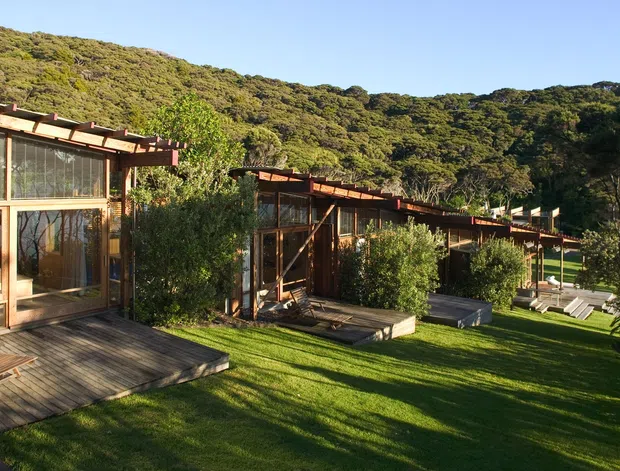 Exterior view of a low-rise timber-clad building composed of several connected or repeating pavilion-like volumes, with broad glazed walls, projecting pergola-like roof structures, and timber decks opening onto a manicured lawn. Dense bush-covered hills form the immediate backdrop, suggesting a secluded natural site. Warm late-day sunlight emphasizes the timber structure and greenery.