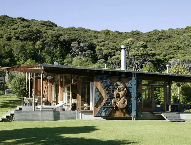 Single-storey low-profile house set in a lush, wooded landscape. The building has extensive timber-framed glazing, a broad shallow roof, dark horizontal roof edging, timber cladding, and outdoor deck areas. A prominent geometric timber feature wall with sculptural carved elements forms the centre of the facade. The surrounding lawn and dense native vegetation are visible, with multiple metal flues projecting through the roof.