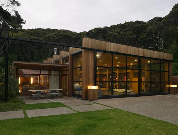 Exterior view of a low-profile contemporary residence set within dense native forest. The home features extensive floor-to-ceiling glazing, warm vertical timber cladding, a projecting flat roof, and a covered outdoor living and dining area connected to the main volume. Concrete stepping slabs, lawn, and a paved driveway frame the building.