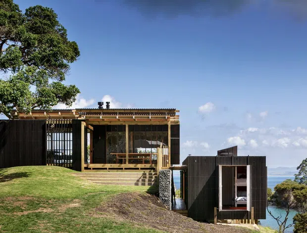 Exterior view of a compact modern coastal residence on a sloping grassy site, composed of dark corrugated-clad volumes with timber-framed glazing and a raised deck. The main pavilion has a low pitched roof and screened outdoor living area, while a smaller lower-level volume projects toward the ocean. Mature trees and a visible coastline form the surrounding setting.