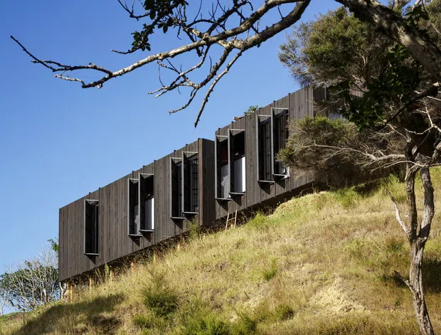 Exterior view of a long, narrow building set into a steep grassy, wooded hillside. The facade is clad in dark weathered vertical timber boards and punctuated by a regular series of tall projecting window bays with dark frames. The building appears elevated or stepped along the slope, with a strong linear form and minimalist detailing under a clear blue sky.