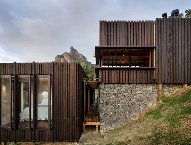 Exterior view of a contemporary hillside dwelling composed of dark vertical timber slat walls, extensive glazing, and a rugged stone retaining wall. A partially enclosed upper-level living space opens toward a dramatic rocky mountain peak in the background, while a narrow timber walkway connects the building volumes.