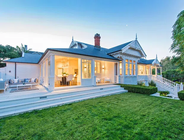 Exterior view of a refined single-storey heritage-style house with a pale weatherboard facade, ornate gabled roof details, decorative timber trim, a brick chimney, and a broad tiered veranda and deck. Large glazed doors reveal a warmly lit dining and living area, while landscaped hedges and a lawn surround the home. An outdoor lounge is visible on the left side of the deck.