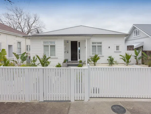Street-facing view of a compact single-storey white weatherboard home with a low-pitched metal roof, symmetrical facade, louvred windows, dark entry door, and a white vertical-slat front fence enclosing a small planted garden. Neighbouring weatherboard homes are visible on either side.