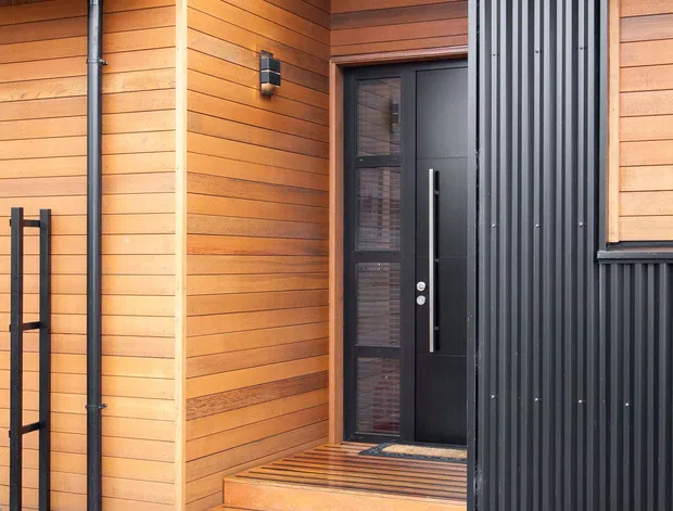 Close-up exterior view of a modern timber-clad building entrance. Warm horizontal wood weatherboards meet a contrasting black vertical corrugated metal facade, with a black-framed glazed entry door, covered porch, timber steps and deck, and a gravel stepping-stone path bordered by rocks and planting. The roof has dark metal roofing and exposed timber eaves.