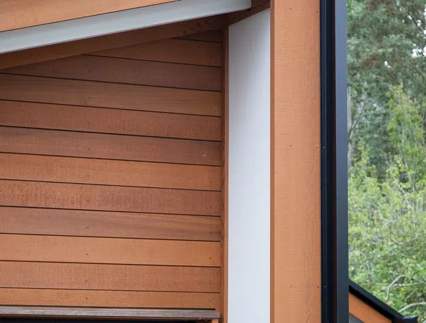 Close-up exterior architectural detail showing a dark metal-framed opening with warm horizontal timber cladding and timber structural trim. A black garage or door panel is visible at the lower left, with a white recessed wall surface and a pitched roof edge beyond. Trees form the background.