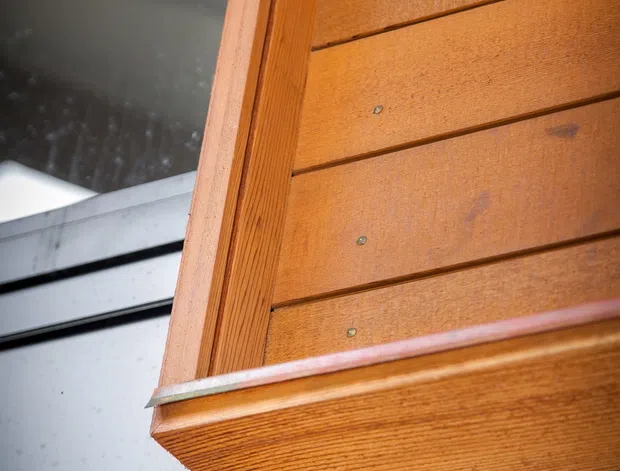 Close-up architectural detail of warm-toned horizontal timber cladding and a projecting timber edge or soffit, with visible metal fixings and dark metal framing or roofing below. The image focuses on exterior construction materials and joinery rather than a complete room or building.
