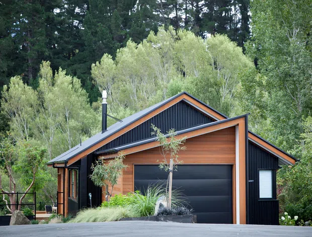 Exterior view of a compact contemporary home or garage-fronted residence set among mature trees. The building has a steep gabled roof, dark vertical cladding, warm timber-toned horizontal wall panels, a large black sectional garage door, and a visible metal flue. Landscaping partially screens the lower facade.