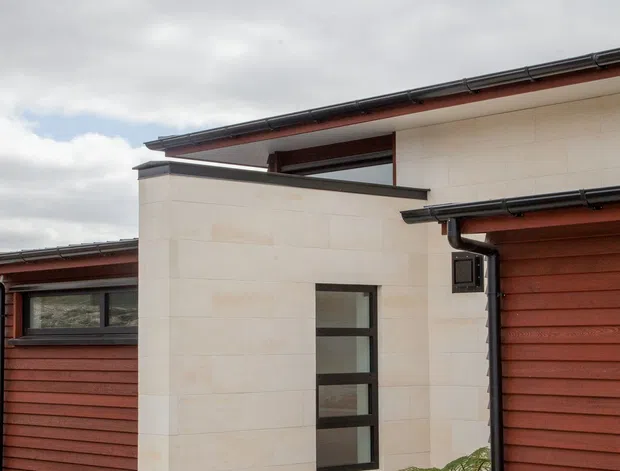 Exterior view of a contemporary multi-level residential building with pale masonry wall sections, horizontal reddish-brown timber cladding, dark metal roof edging and gutters, and narrow black-framed windows. A concrete walkway and minimalist dry-rock planting bed with small palms frame the facade.