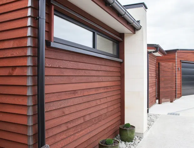 Exterior view of a contemporary residential building featuring horizontal red-brown timber cladding, dark metal gutters and downpipes, black-framed windows, pale masonry panels, and a clean concrete walkway bordered by white decorative stones and potted plants.