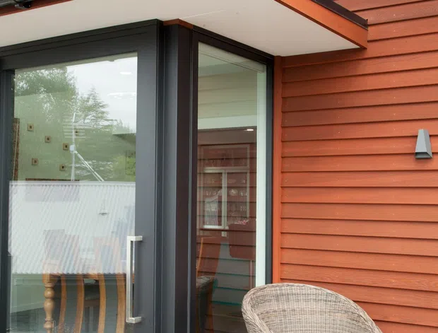 Close-up exterior view of a modern timber-clad house entrance opening onto a covered timber deck. The facade has warm reddish-brown horizontal weatherboard cladding, black-framed full-height glazed doors, exposed gutters and downpipes, and a simple projecting canopy. A wicker outdoor chair and small timber side table sit by the entry.