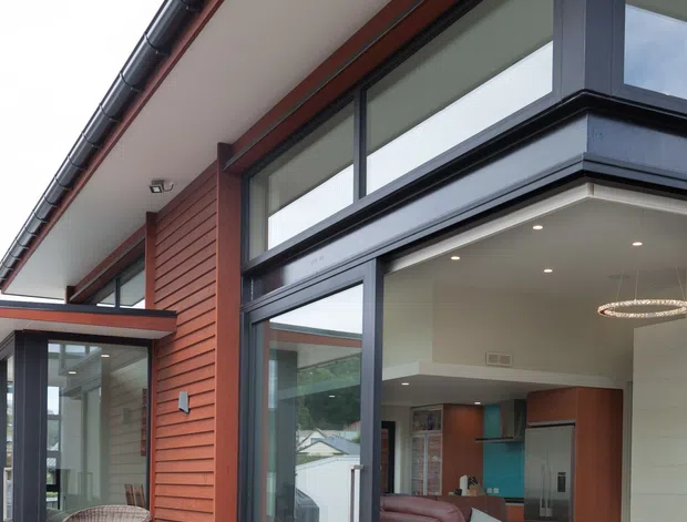 Contemporary residential exterior viewed from a timber deck, with red horizontal cladding, dark metal-framed floor-to-ceiling glazing, and a covered roof edge with gutters. The open glazed doors reveal a modern living room with a leather recliner, rug, built-in cabinetry, and a kitchen area beyond. A covered outdoor seating nook is visible to the left.