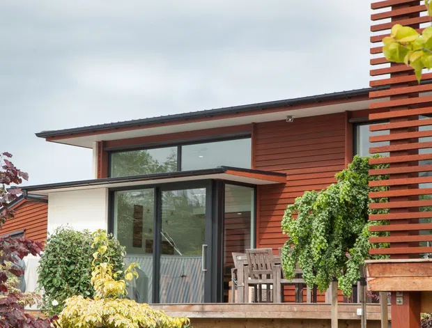 Exterior view of a contemporary single-storey home with a low-pitched roof, extensive dark-framed glazing, warm horizontal timber cladding, and a small raised timber deck. A vertical timber privacy screen and established planting frame the entrance and outdoor seating area.