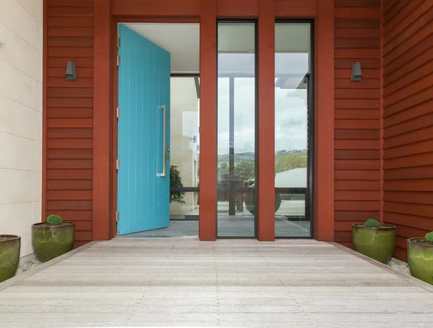 Exterior entry viewed from a timber deck, featuring a vivid turquoise front door, tall timber-framed glazed panels, red-brown horizontal timber cladding, pale stone wall lining, and potted plants. The glazing reveals a bright interior and distant landscape beyond.