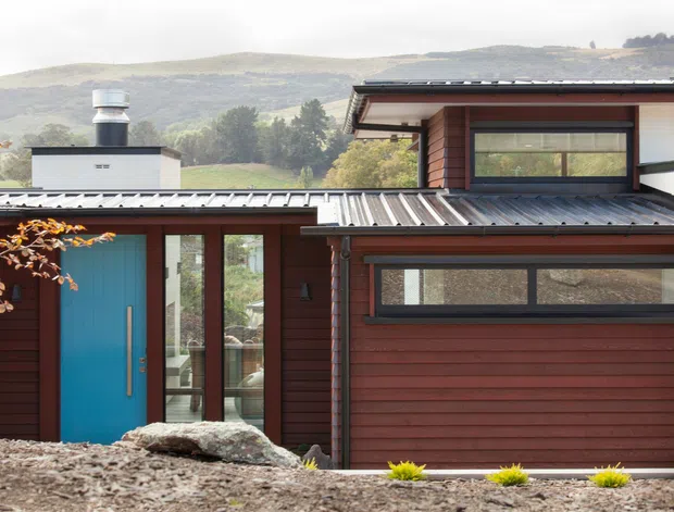 Exterior view of a contemporary single-storey home in a hilly landscape, featuring dark red-brown horizontal cladding, a bright blue entry door, broad horizontal windows, and a low-pitched corrugated metal roof. A garage door is visible at the front, with gravel landscaping and stone accents in the foreground.