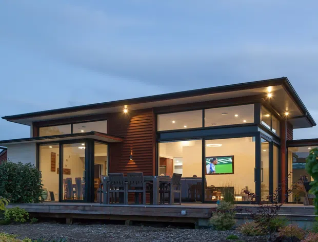 Contemporary single-storey home viewed from the rear at dusk, featuring broad floor-to-ceiling glazing, dark horizontal timber cladding, a low-pitched projecting roof, and an elevated timber deck with outdoor dining furniture. Warm interior lighting reveals an open living area through the windows.