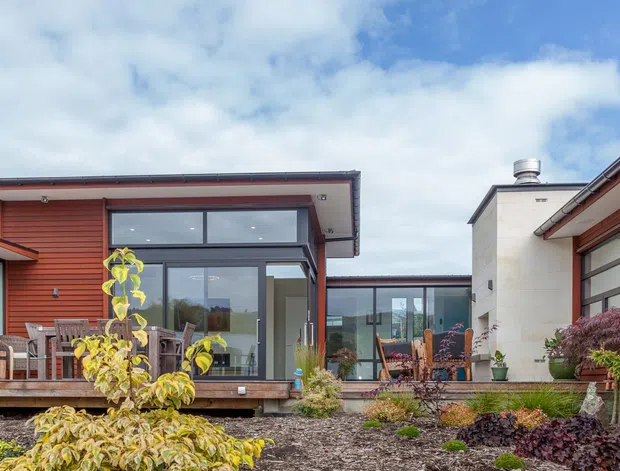 Exterior view of a low-profile contemporary residential home arranged around a landscaped courtyard. The house features red-brown horizontal weatherboard cladding, broad dark-framed glazing, shallow roof overhangs, a timber deck with outdoor dining furniture, and a contrasting light rendered volume. The foreground contains ornamental planting and garden beds beneath a partly cloudy sky.