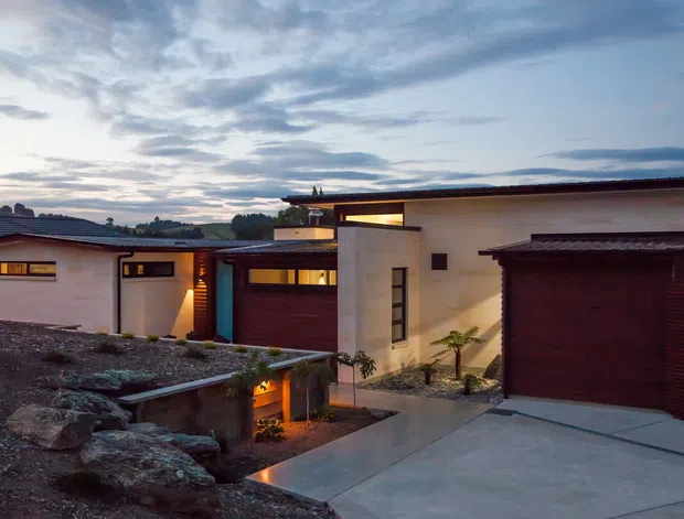 A low-profile modern residential exterior at dusk, arranged across a sloping rocky site. The home features smooth white rendered walls, extensive dark timber garage and entry doors, warm recessed lighting, narrow horizontal windows, and layered flat rooflines. Concrete driveways and paths connect the volumes, with sparse landscaping integrated among exposed rocks. A distant suburban landscape and dramatic cloudy sky form the background.