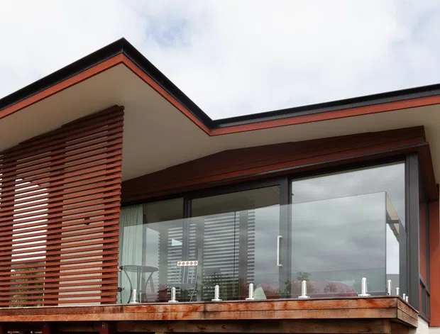 Exterior view of a contemporary single-storey home with a strongly projecting flat roof, broad glazed openings, a timber-lined facade, and a raised balcony or deck enclosed by clear glass. A prominent vertical timber louvre screen provides solar shading and privacy, while dark framing and warm reddish-brown timber accents contrast with the pale roof soffit.