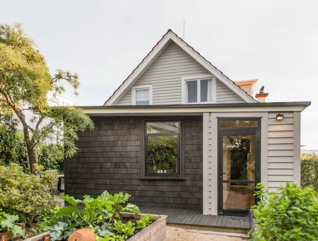 A compact coastal-style detached house viewed from the garden, featuring a steep front gable, light horizontal weatherboard cladding, a dark shingle-clad ground-floor extension, dark-framed glazing, and a small planted garden in the foreground.