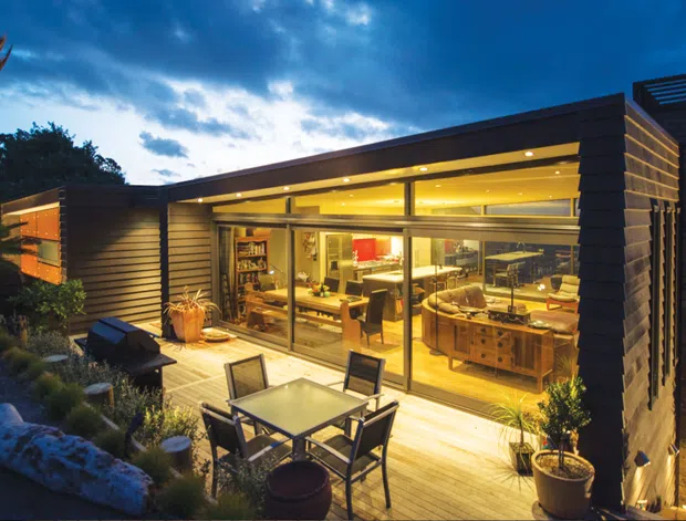 A low-profile contemporary coastal home viewed from an outdoor terrace at dusk. The building has dark horizontal cladding, extensive floor-to-ceiling glazing, warm interior lighting, and a timber deck furnished with an outdoor dining table and chairs. The interior appears to include an open-plan kitchen and living area, while ocean or coastal water is visible beyond the house.